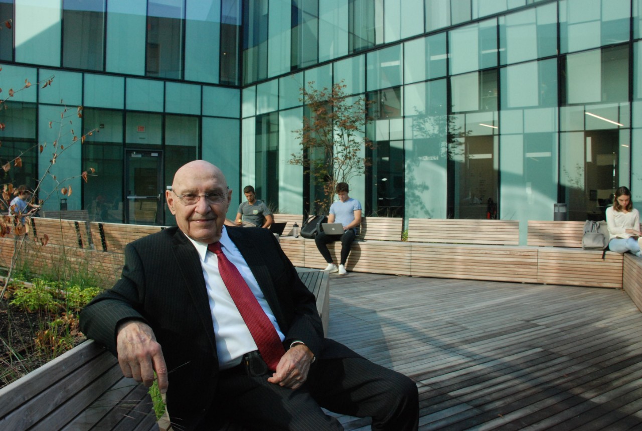 John Goering in a dark suit, white shirt and red tie sits on a bench in front of Lindner College of Business