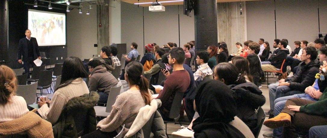 Man stands and speaks in front of a seated audience.