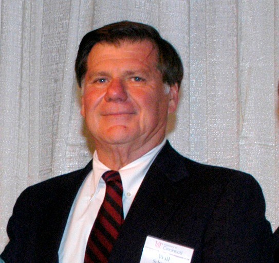 A man in a black jacket and white shirt stands in front of a gray background.