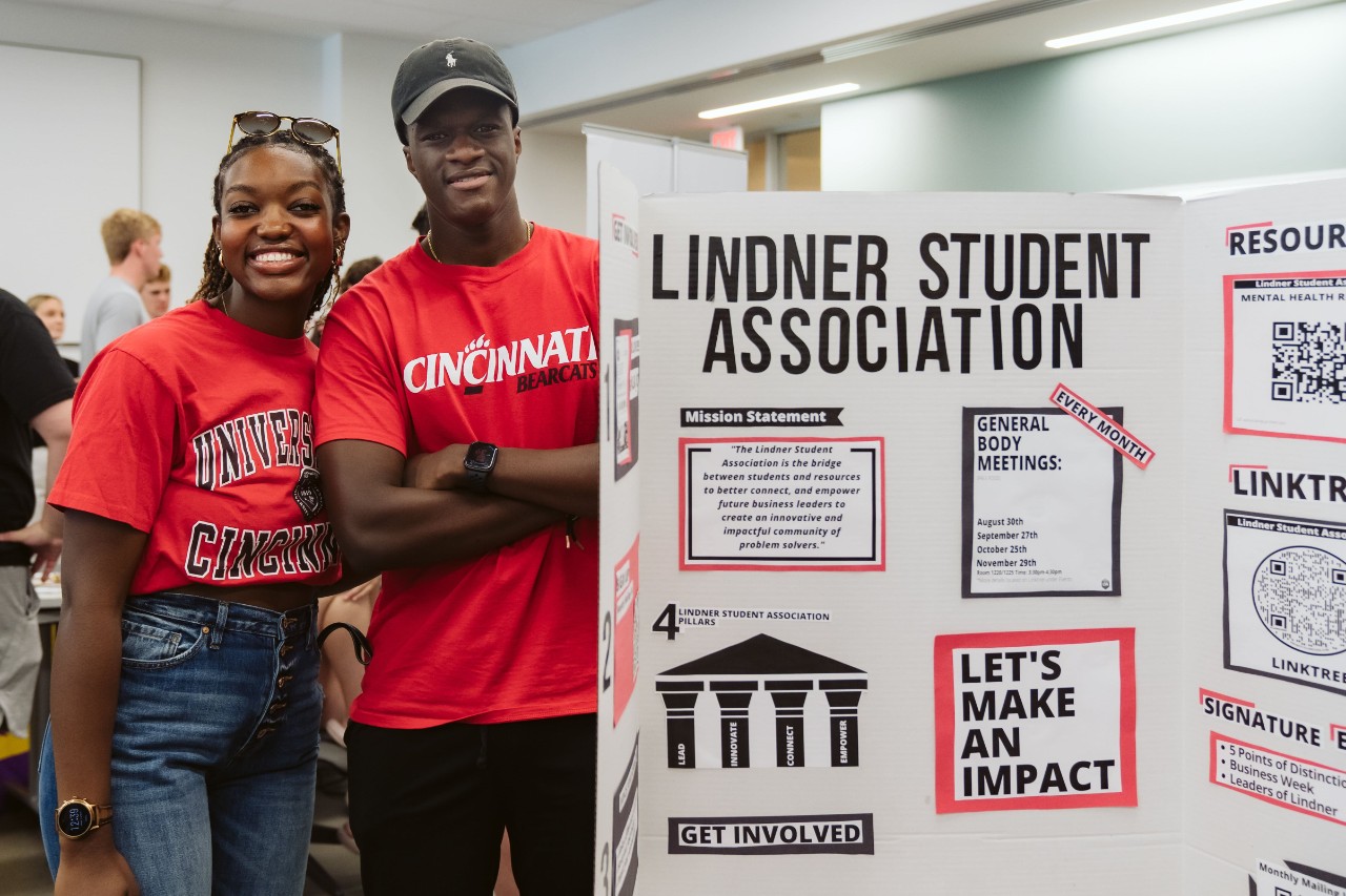 Two students in red tops stand next to a posterboard.