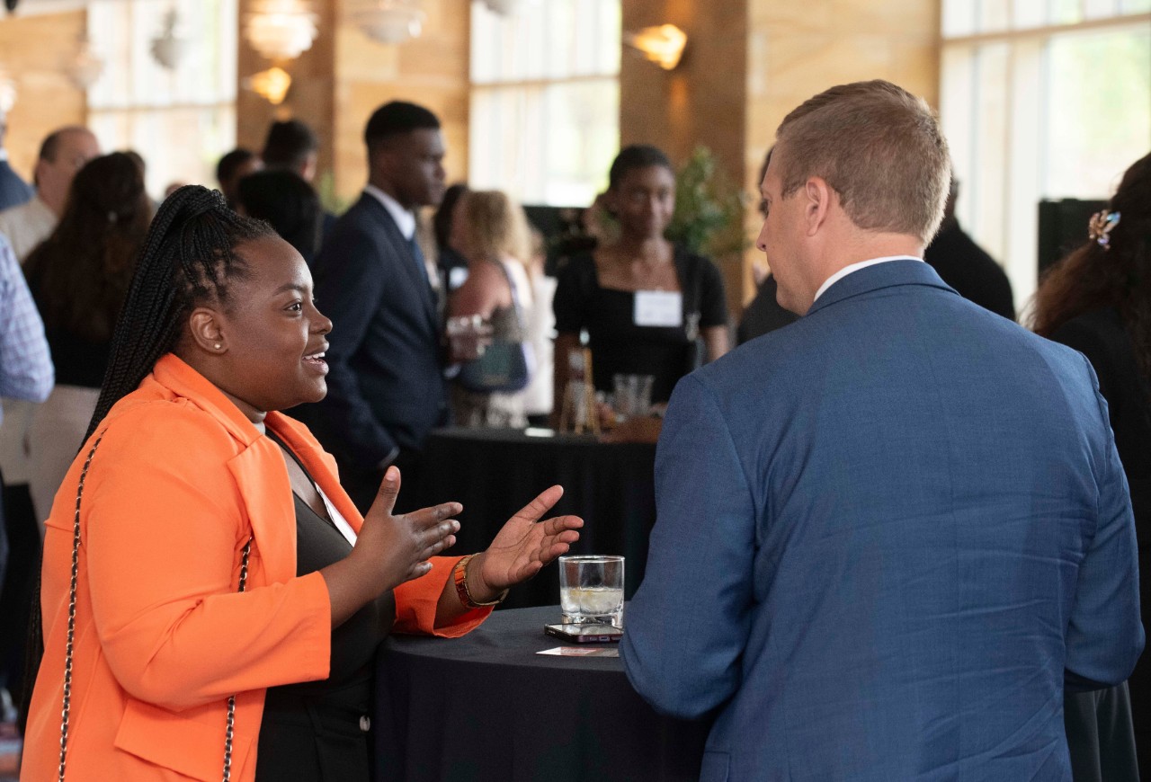 A student in an orange jacket and black shirt talks to a man in professional dress.