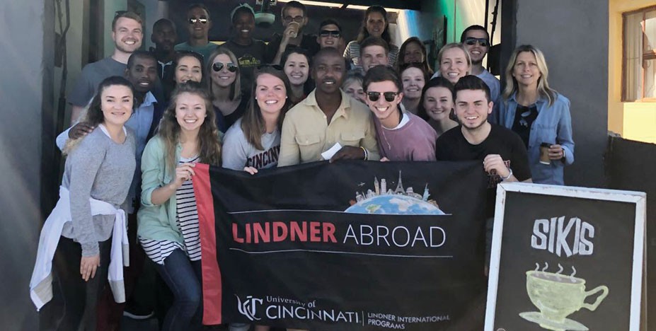 Lindner students, faculty, and staff pose with a coffee shop owner in South Africa, holding a "Lindner Abroad" flag