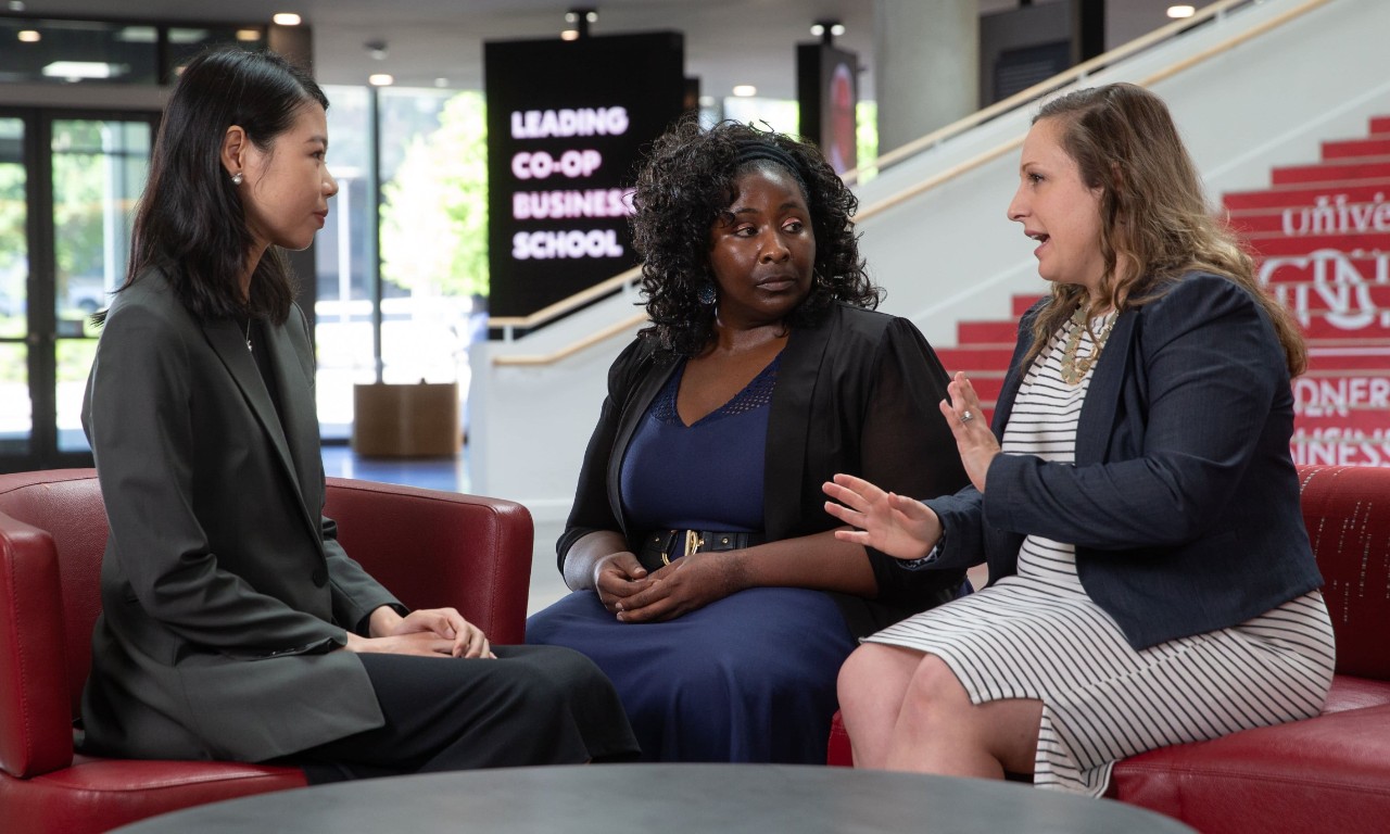 Three female researchers in professional dress talk in the Lindner Hall atrium.