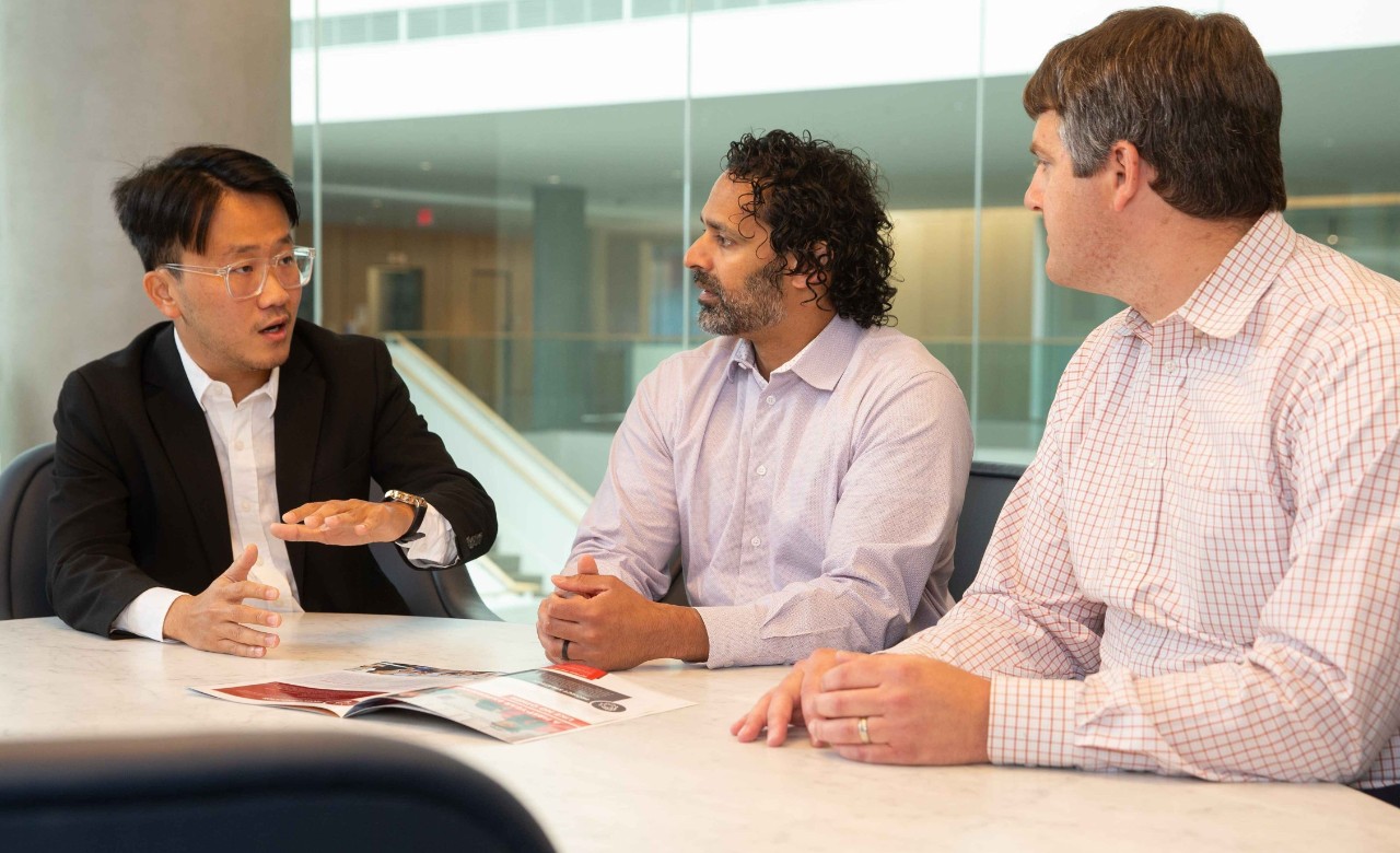 Three men in professional dress sit at a table and talk about their research.