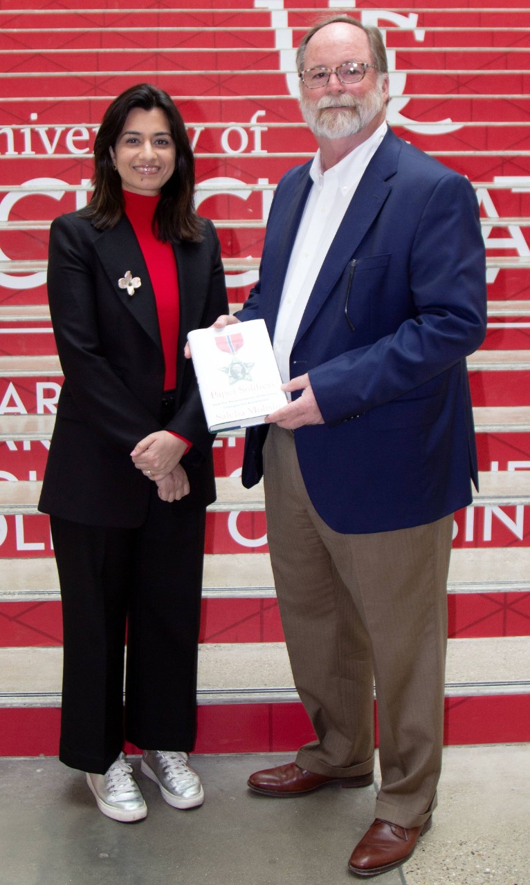 A woman in a black suit and red shirt stand with a man in a blue shirt and brown pants, holding a book.