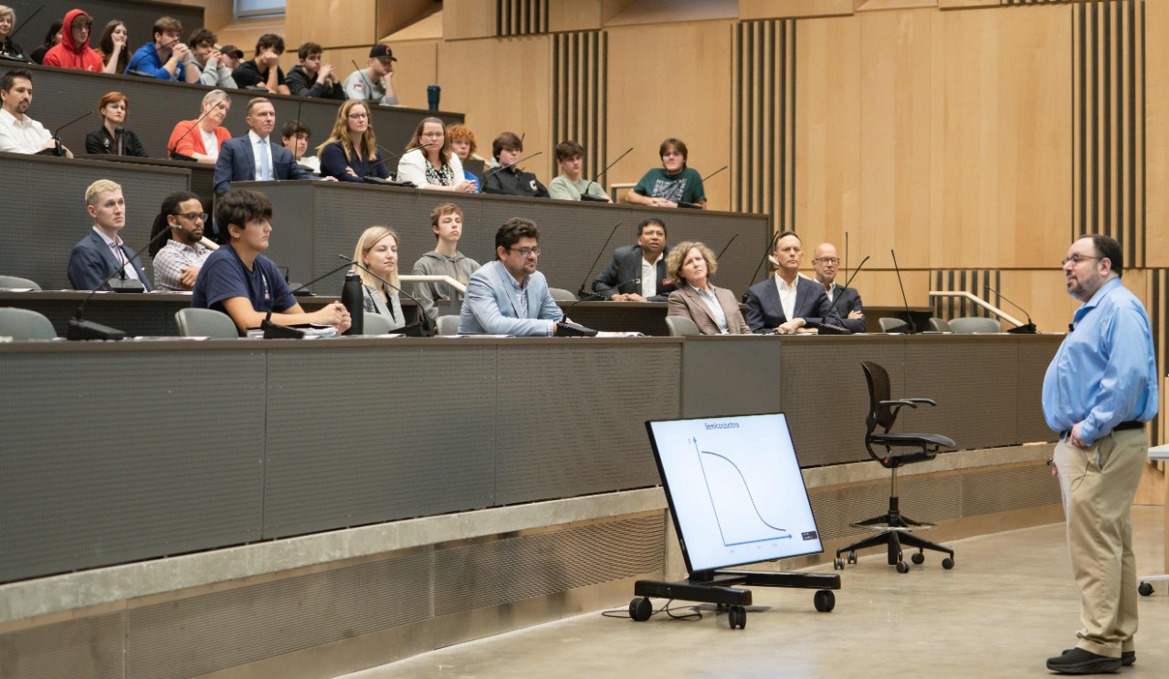 A man in a blue shirt and brown pants speaks to a crowd of economics students and faculty.