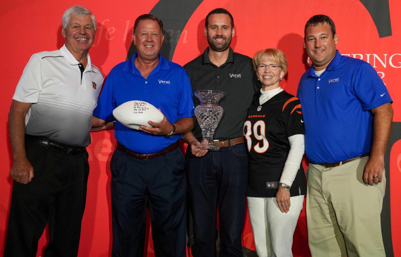 Four men and one woman hold various prizes and stand in front of a red banner.