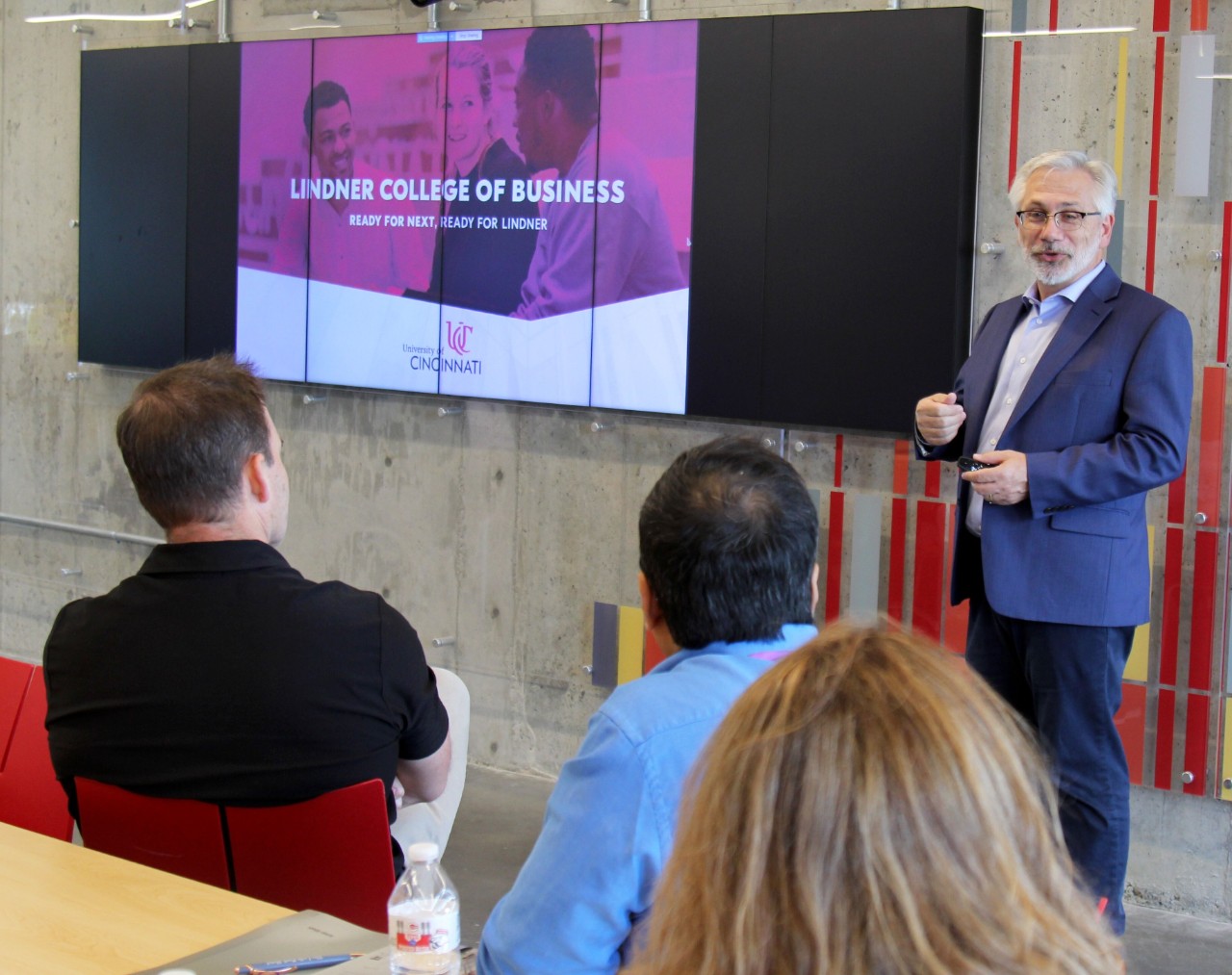 A man in a blue suit stands in front of a TV and instructs of a group of people.