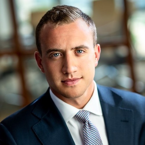 A man with short dark blonde hair smiles for a headshot wearing a white collared shirt, striped tie and navy blue suit jacket