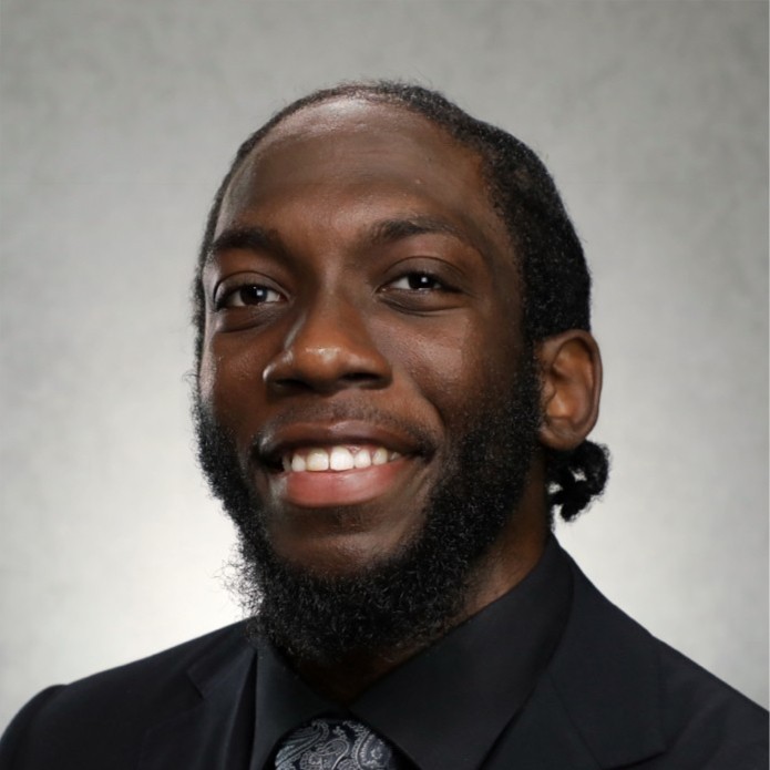 Headshot of Ejiro Agoba in black shirt and jacket in front of grey lit background