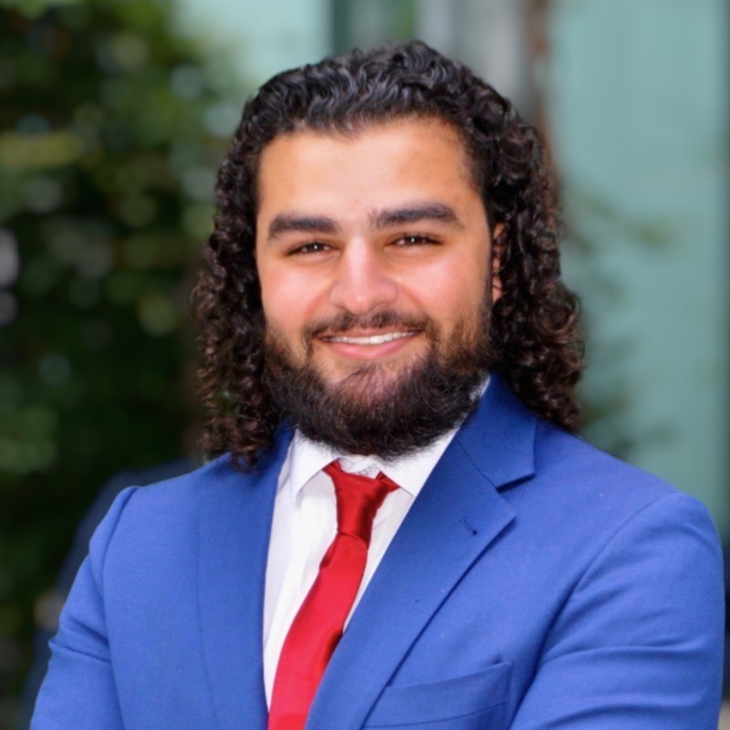 Headshot of Matthew Boutros in a white button-down shirt, red tie, and blue suit jacket in a blurred outdoor environment