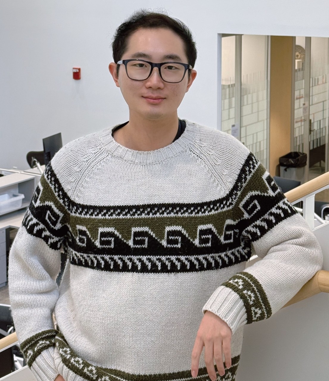 PhD student Zachery Ma poses for a picture on Lindner hall office steps, wearing a white, black and dark green sweater.
