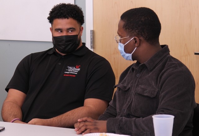 Two young men wearing black tops and face masks sit and face each other in conversation